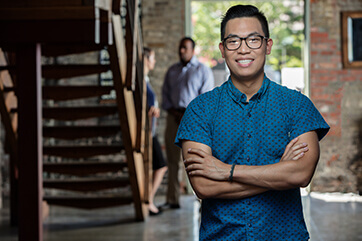 A smiling young man with arms crossed in a bright hallway or atrium with stairs in the background
