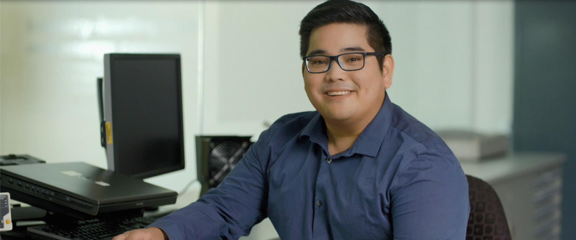 A man working at a desk facing the camera with a monitor in the background