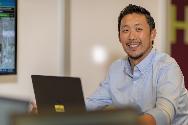 A man with a laptop sitting at his desk smiling