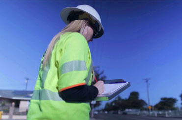 A woman wearing a yellow protective jacket and hard hat writing on a clipboard