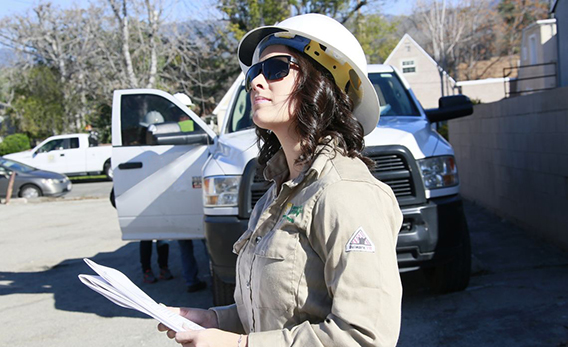 A woman wearing hard hat and sunglasses, has paper in hand and is looking up