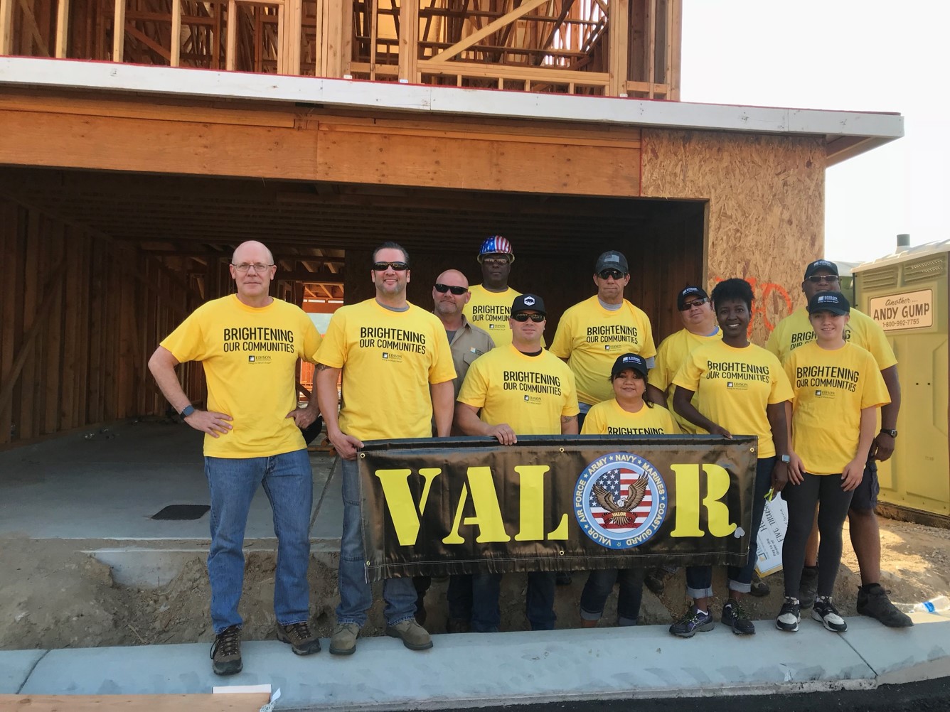 Group of veterans in a home building work site, holding up a sign that says 'Valor', and wearing yellow shirts that say 'Brightening Our Communities'