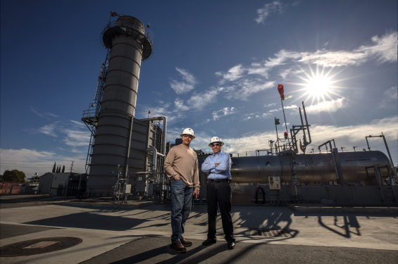 Two men outside at a utility facility wearing hard hats facing the camera