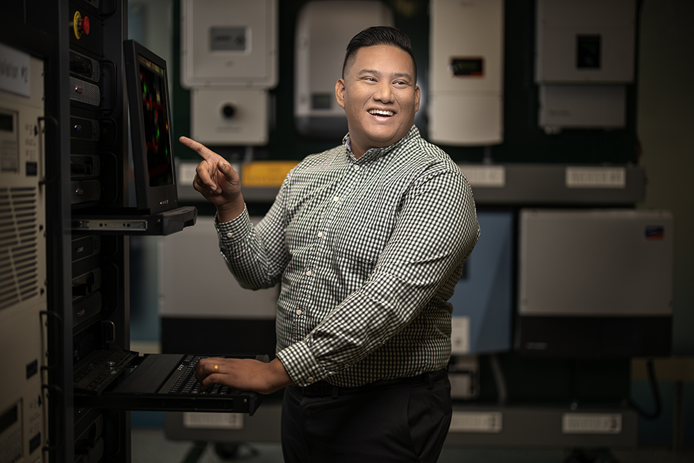Professional engineer standing in front of a computer in an electrical room type environment, smiling and typing on the keyboard and pointing to the computer screen.